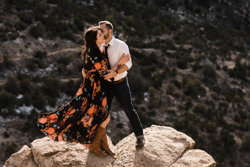 Bride wearing a floral and black wedding dress at her adventurous elopement while the groom kisses her cheek at an overlook on Mount Lemmon.